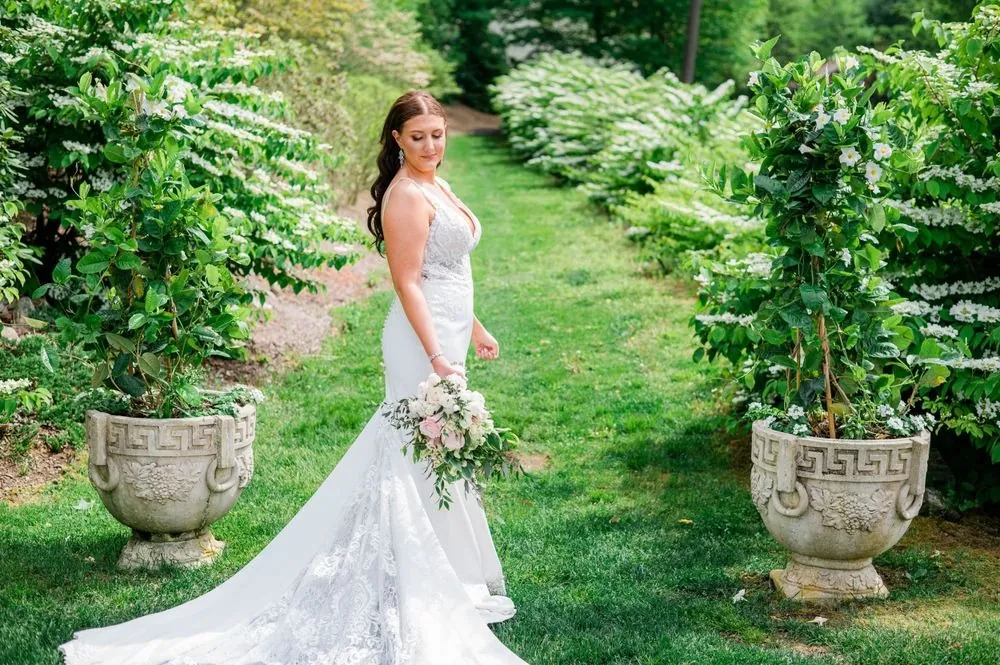 The bride stands on a garden path between potted shrubs, her train spread behind