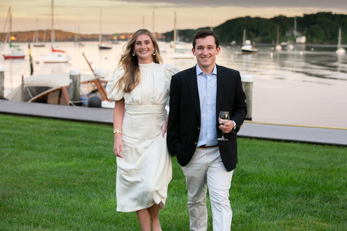 Kate in a cream dress and Parker in a navy blazer on a harborside lawn with moored sailboats