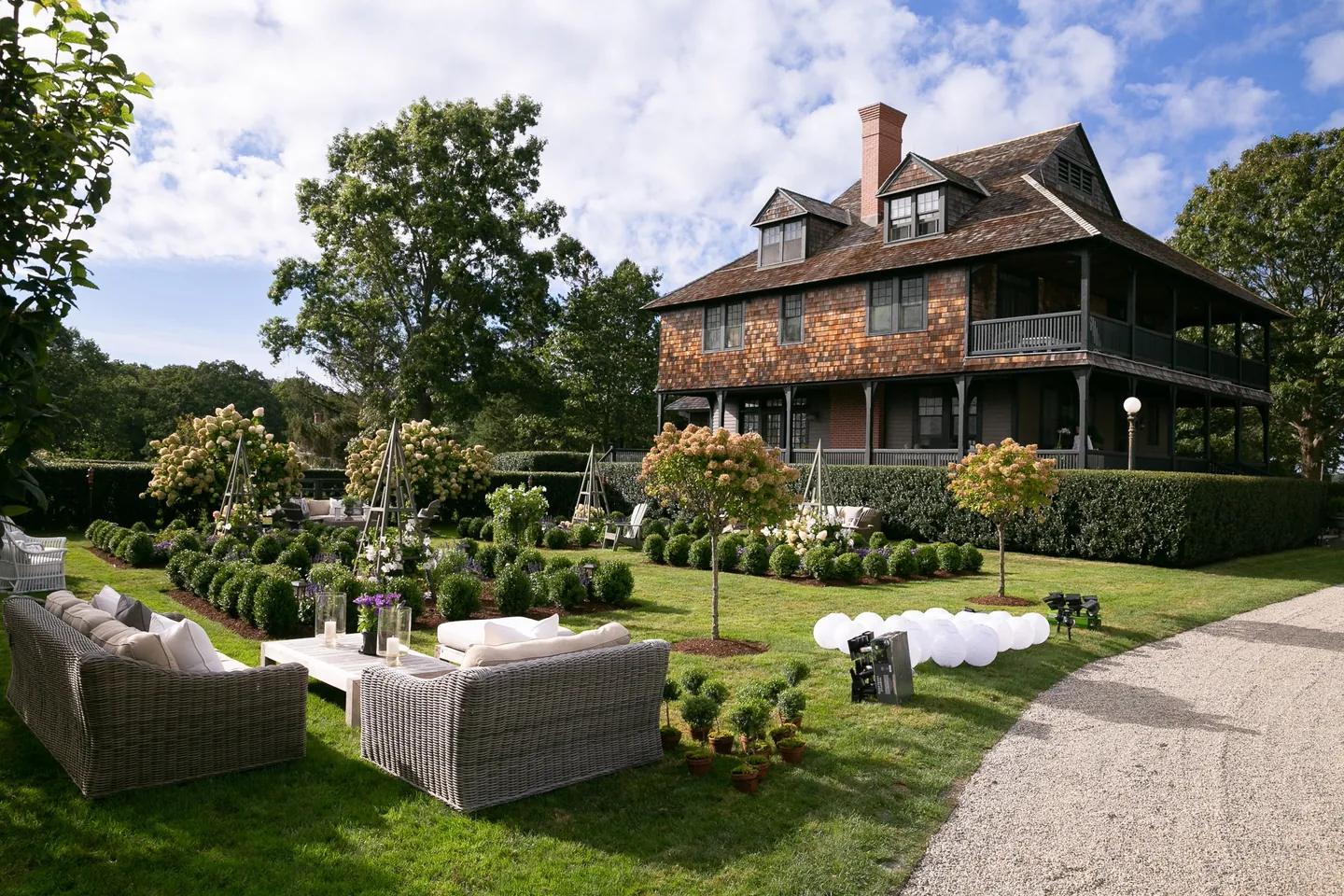 A large shingled house with a wraparound porch and lawn furniture on a sunny afternoon