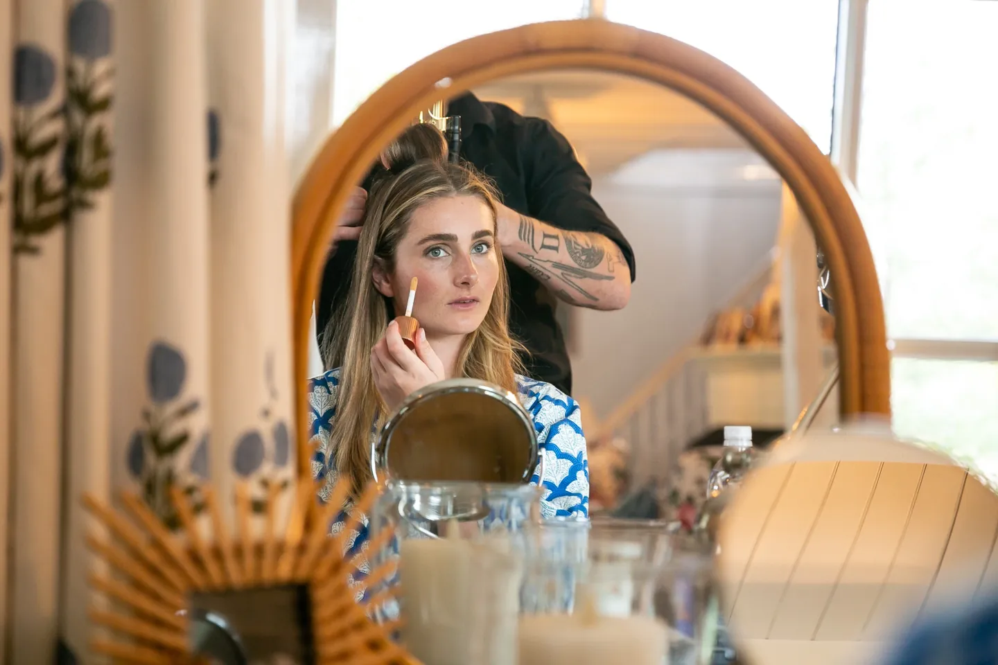 The bride is reflected in a round rattan mirror as her makeup is finished