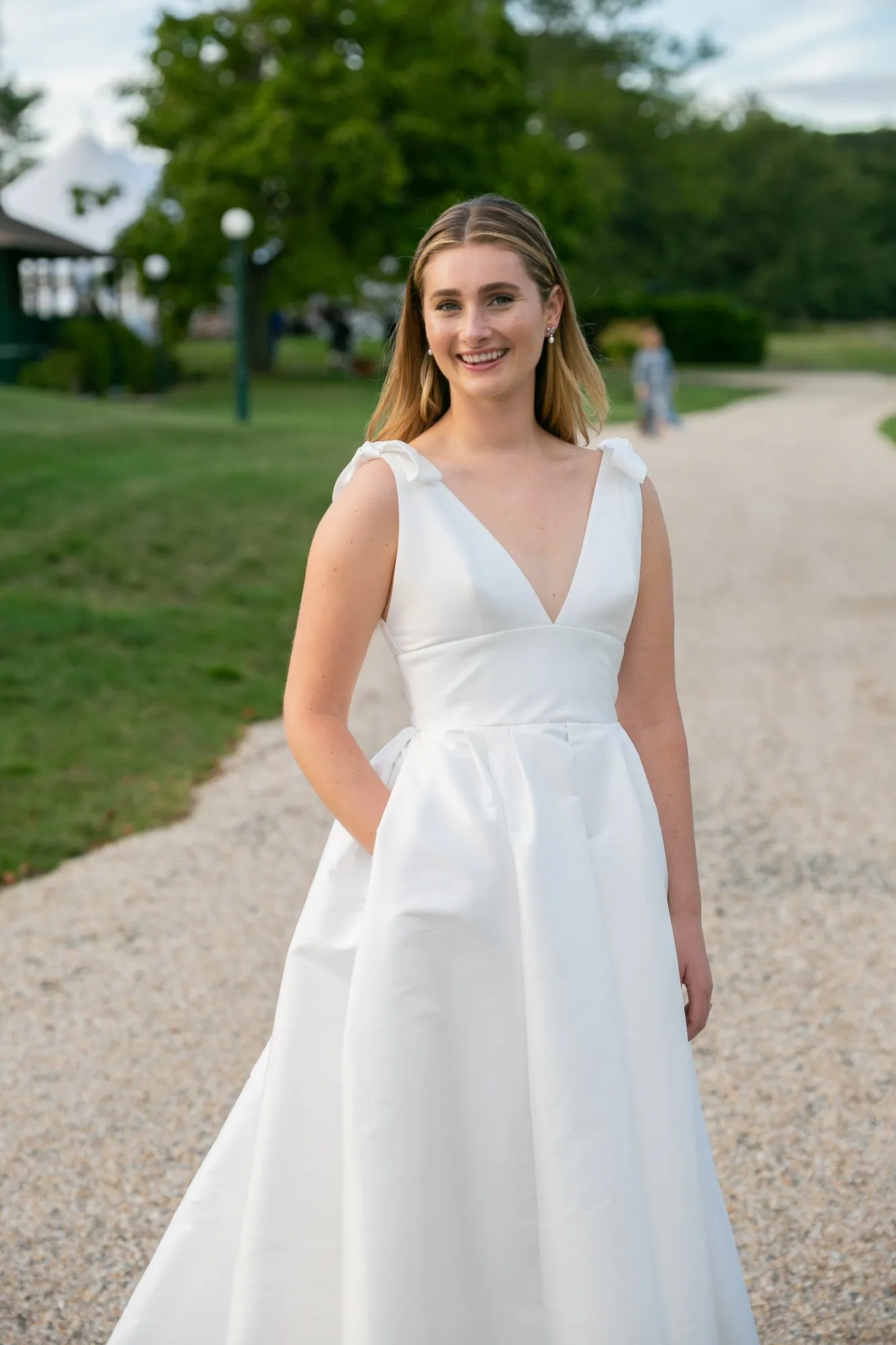The bride stands on a gravel drive in a V-neck gown with bow straps