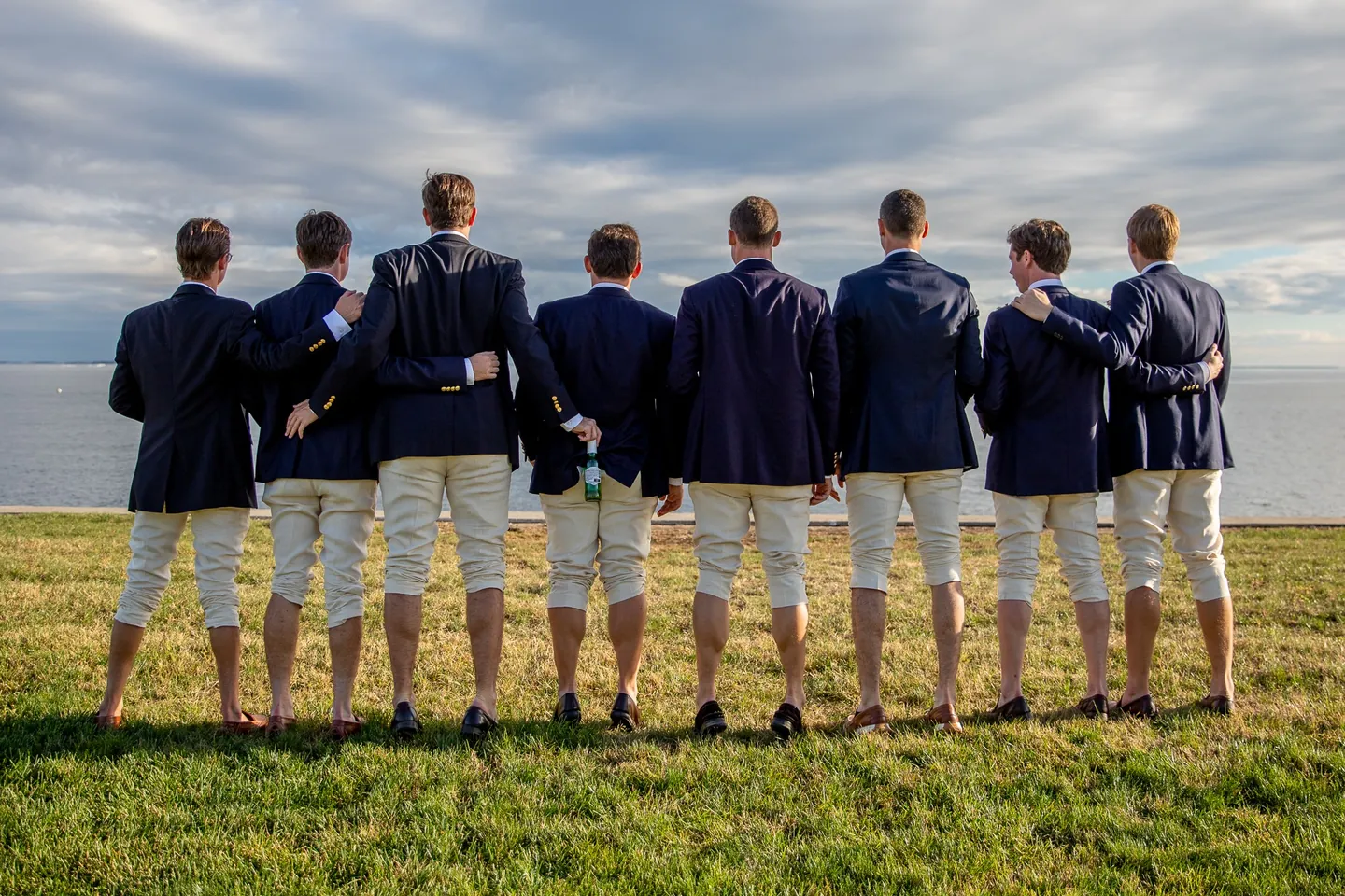 Groomsmen stand shoulder to shoulder with their backs to the camera, facing the water