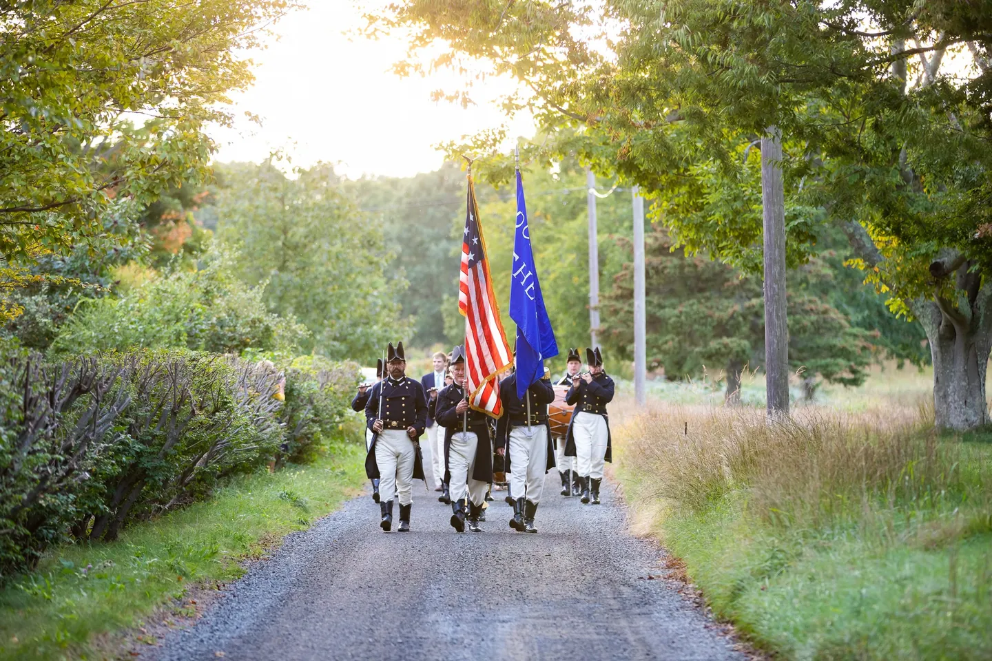 A fife and drum corps in tricorn hats marches down a tree-lined gravel lane under flags