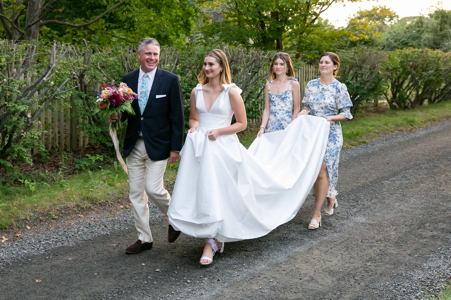 An older man walks the bride down the lane as bridesmaids in toile lift her train
