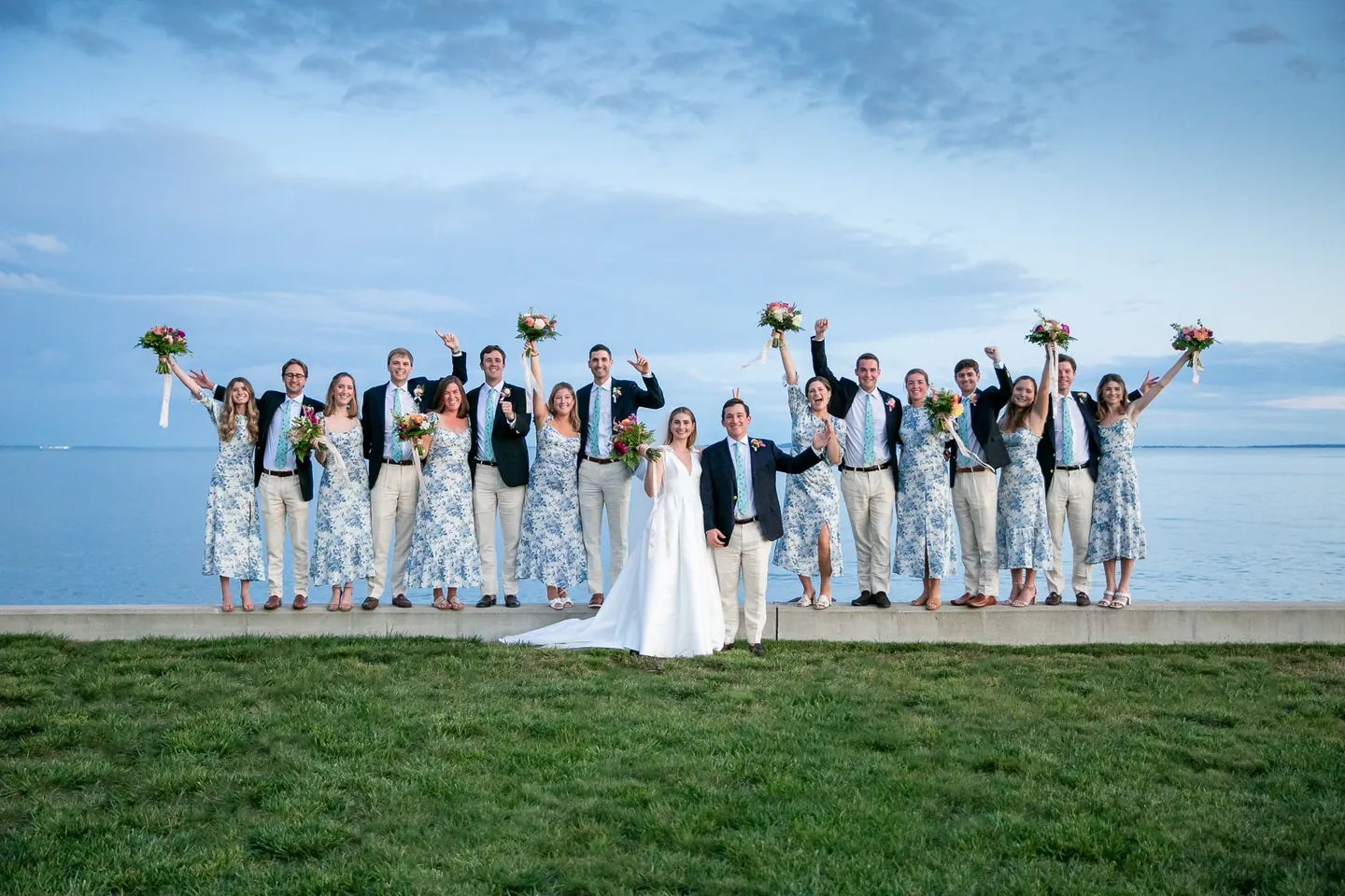 The full wedding party throws bouquets and fists in the air on a seawall above the water