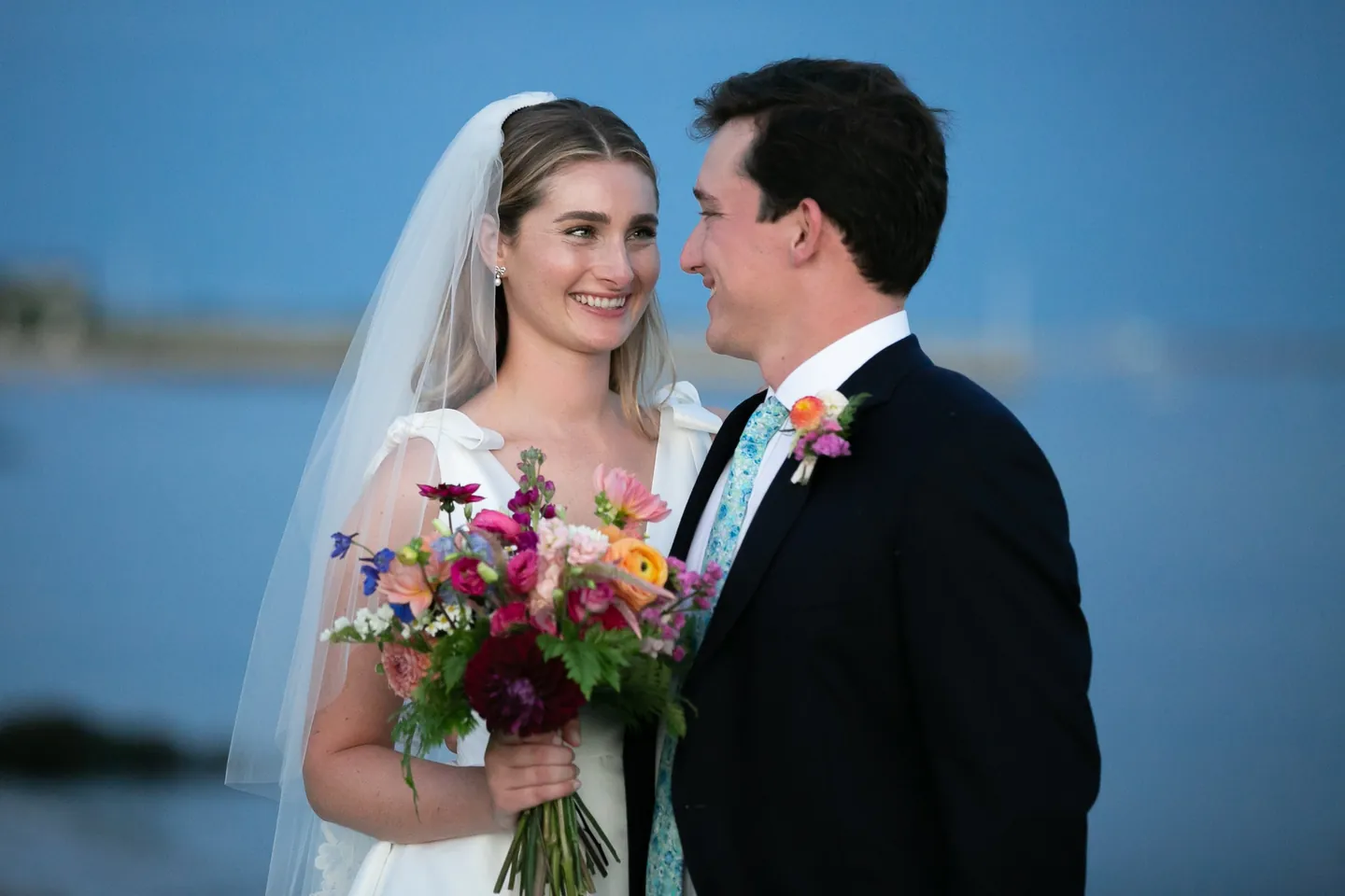 Kate and Parker smile at each other at blue hour, her bright bouquet between them