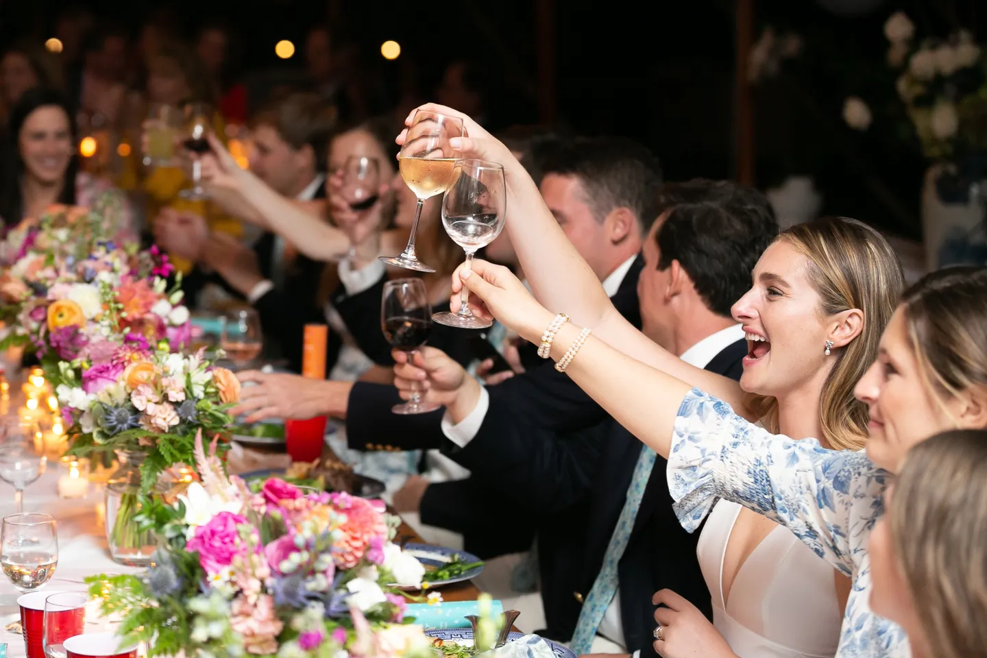 Guests raise glasses and cheer across a flower-covered table at night
