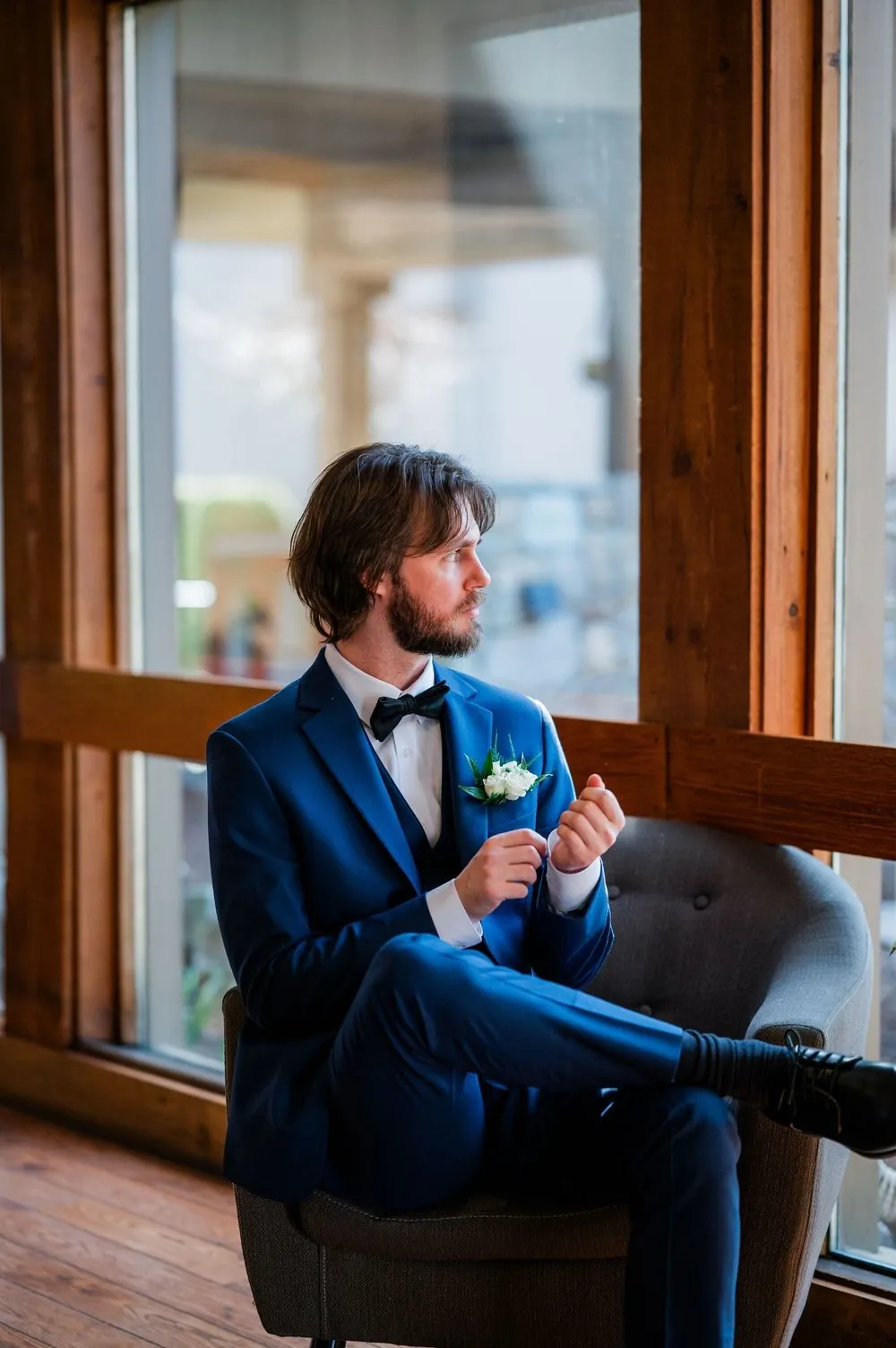 The groom in a cobalt suit adjusts his cuffs in an armchair beside a tall window
