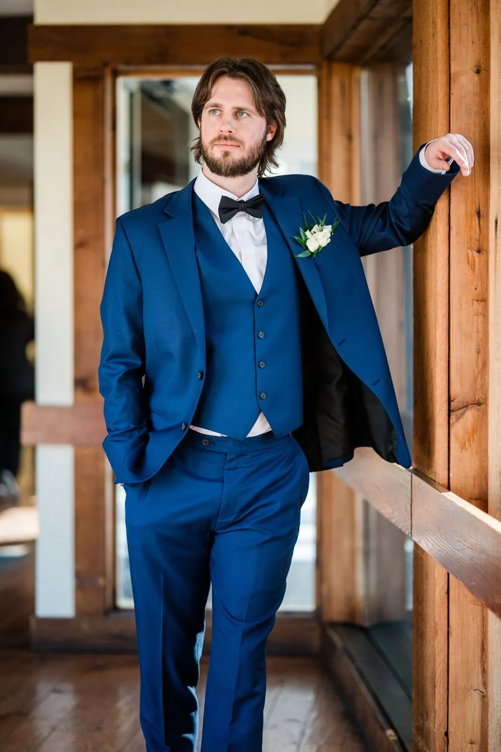 The groom leans against a wood doorframe, one hand on the frame, in blue with a bow tie