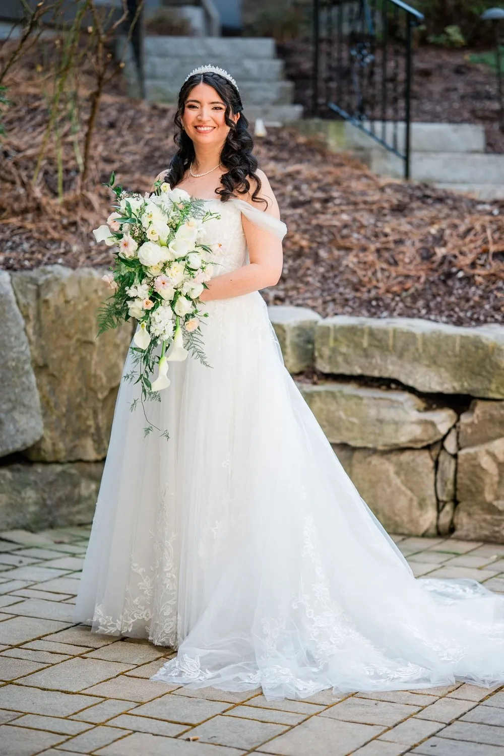 The bride holds a white rose bouquet on a stone path outside the hotel