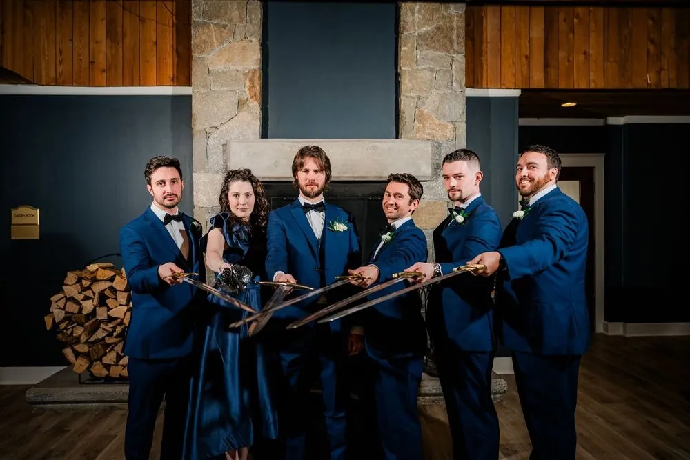 Wedding party in blue holds swords crossed toward the camera before a stone fireplace
