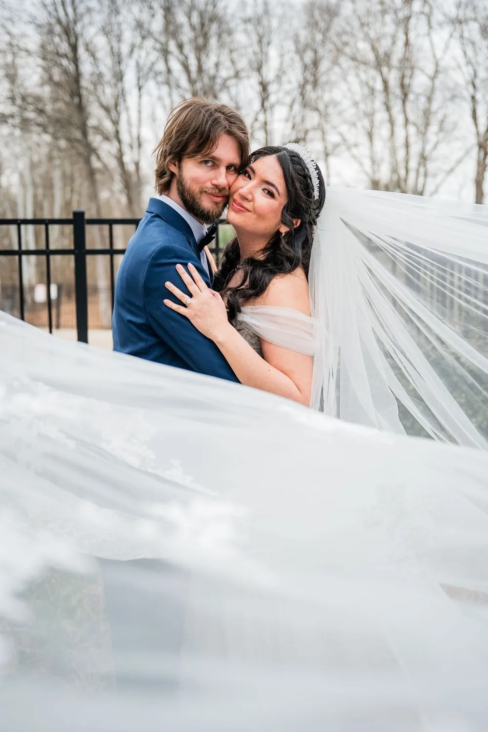 Monica and Kevin lean in close as her long veil sweeps sideways in the wind