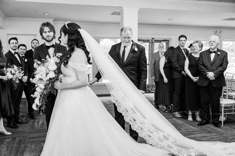 Black and white of the bride brought down the aisle by a man in a tuxedo, her lace veil trailing