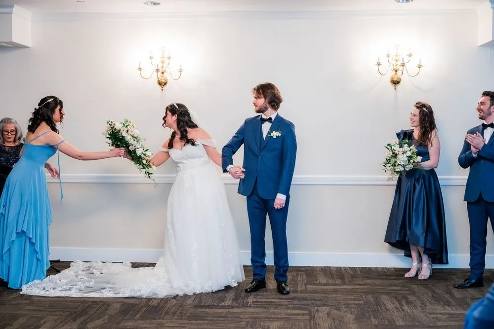 A bridesmaid in blue takes the bride's bouquet at the front of the ceremony room