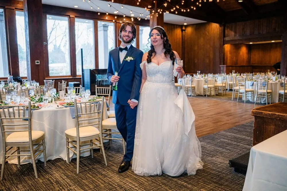 Monica and Kevin walk into the reception hall past set tables and big windows