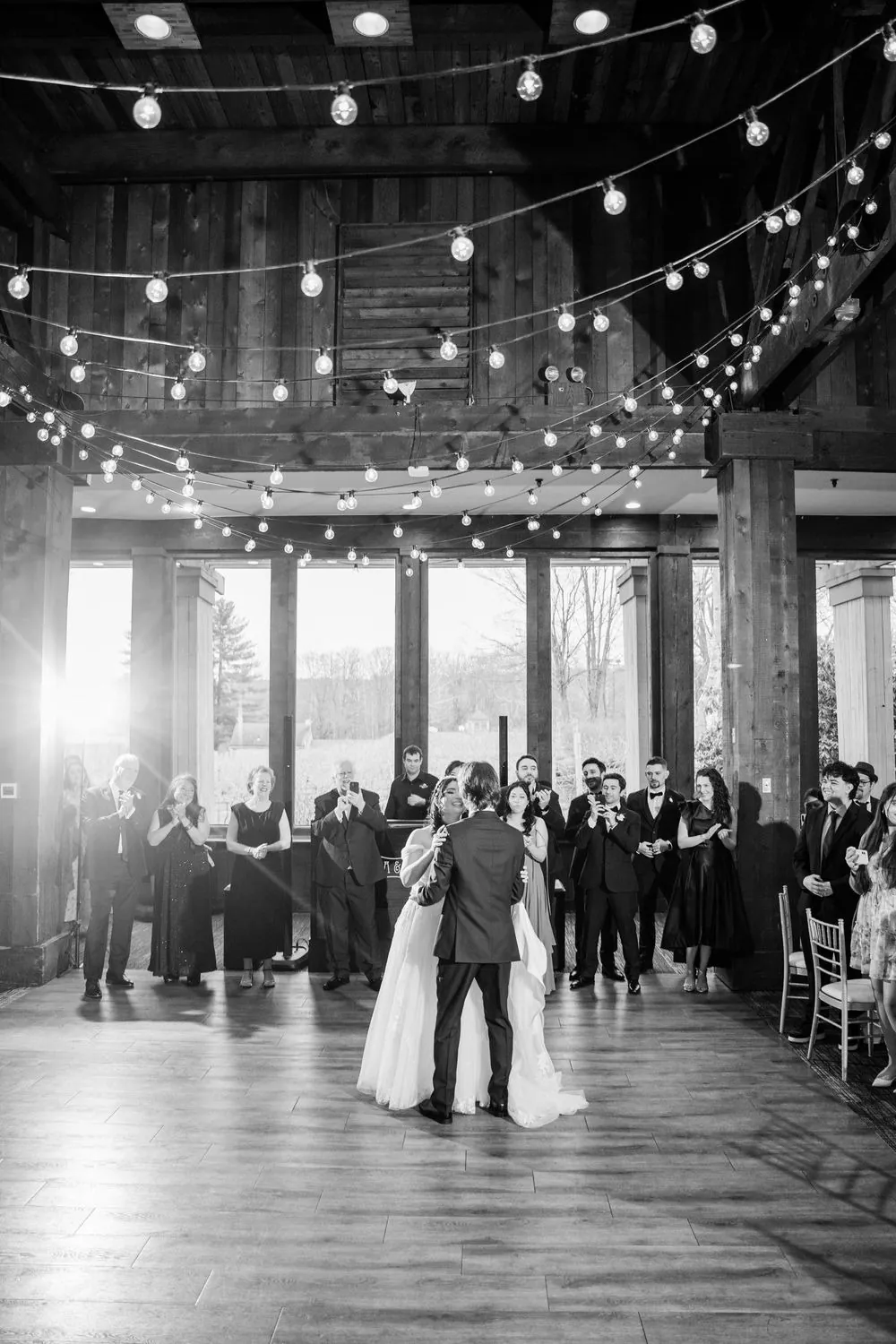 Black and white of the first dance under string lights with guests along the windows