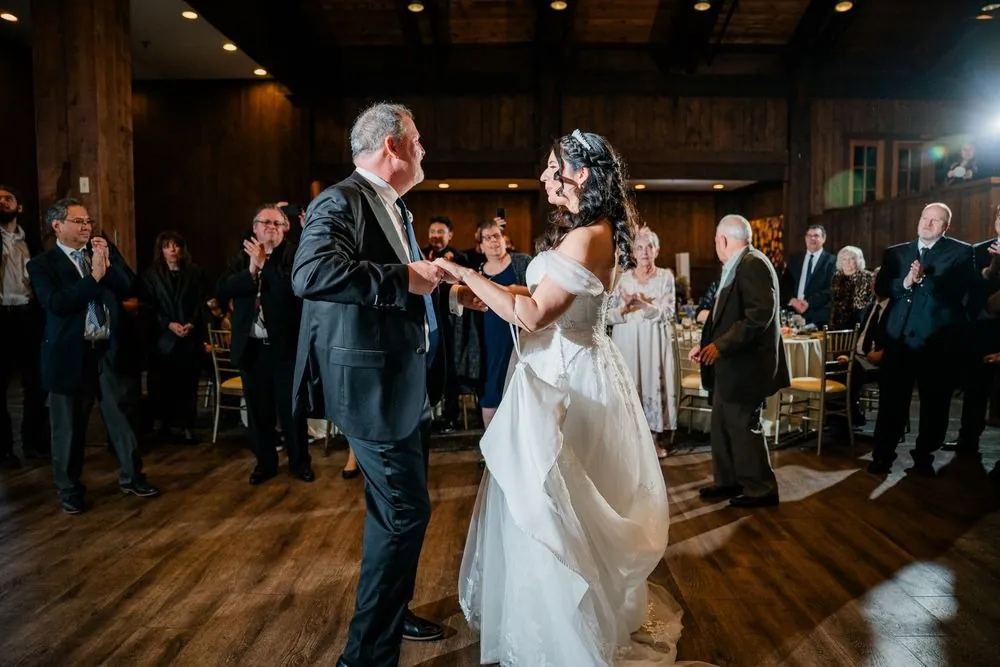 The bride dances with a gray-haired guest in a dark suit as the room claps around them