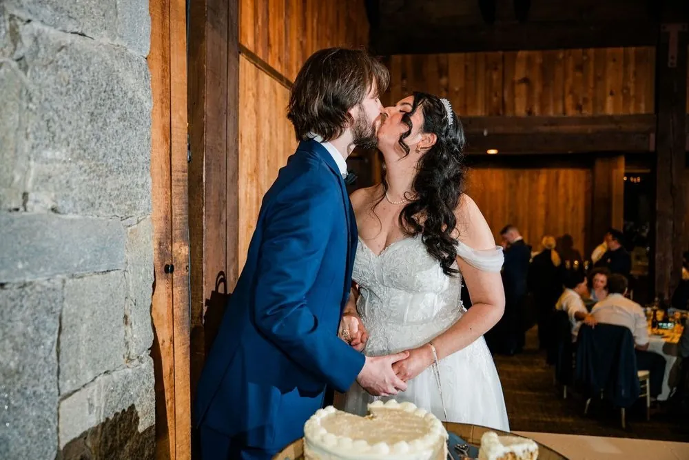 Monica and Kevin kiss beside the cake against a stone wall