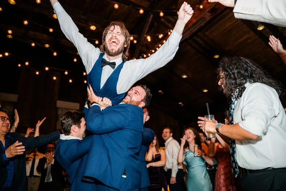 Groomsmen hoist the groom onto their shoulders, his arms flung high under string lights