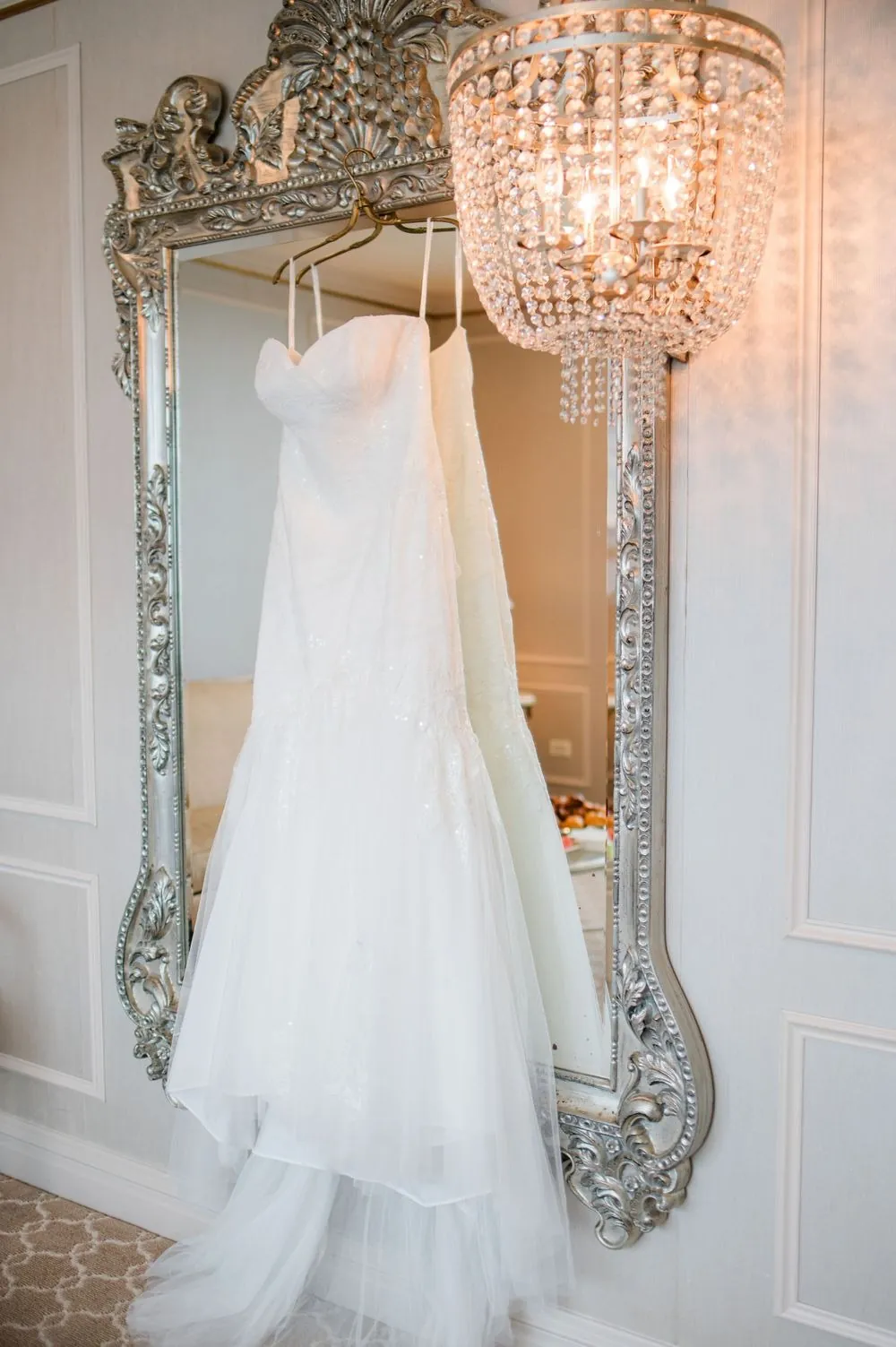 Wedding gown hanging from a tall silvered mirror beneath a crystal chandelier