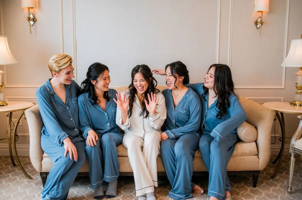 Bridesmaids in dusty blue satin pajamas laugh with the bride on a cream sofa