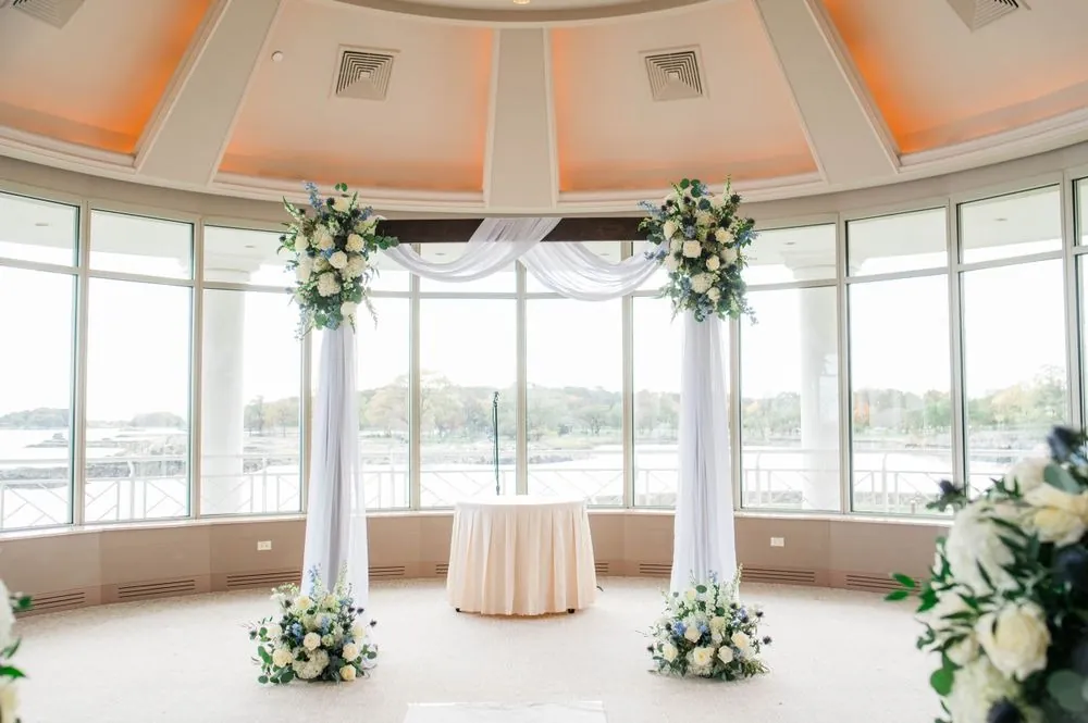 Round ceremony room with a white draped arch, floral arrangements, and water views