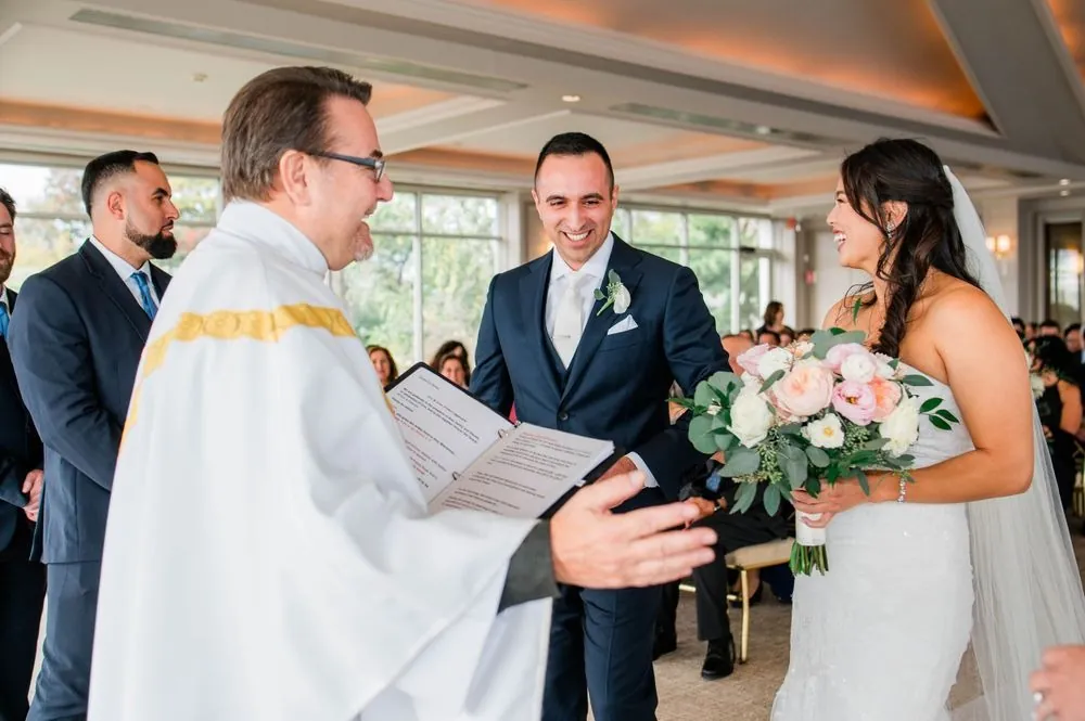 Priest laughs and gestures with his book as the bride and groom grin at the altar