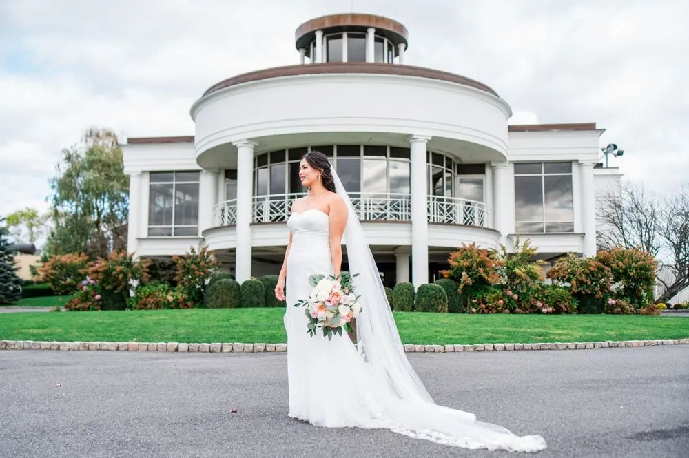 Bride stands on the drive in front of the white columned rotunda with her long veil trailing