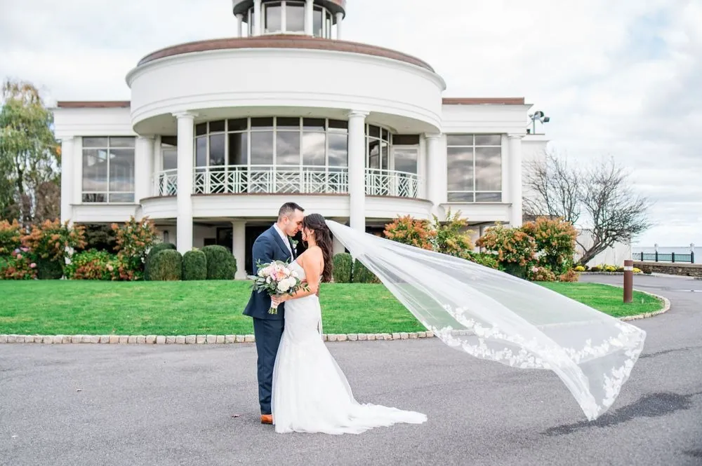 Kim and Fabiano embrace in front of the white rotunda as wind lifts her veil sideways