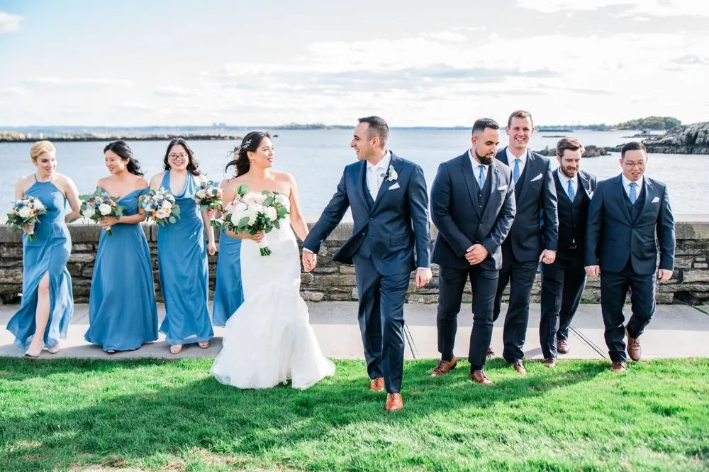 Bridesmaids in dusty blue and groomsmen in navy walk the lawn above the water
