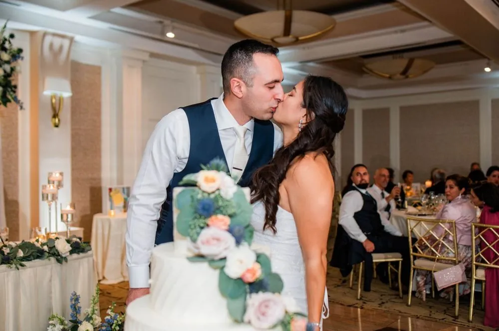 Kim and Fabiano kiss beside a white cake trimmed with pale flowers