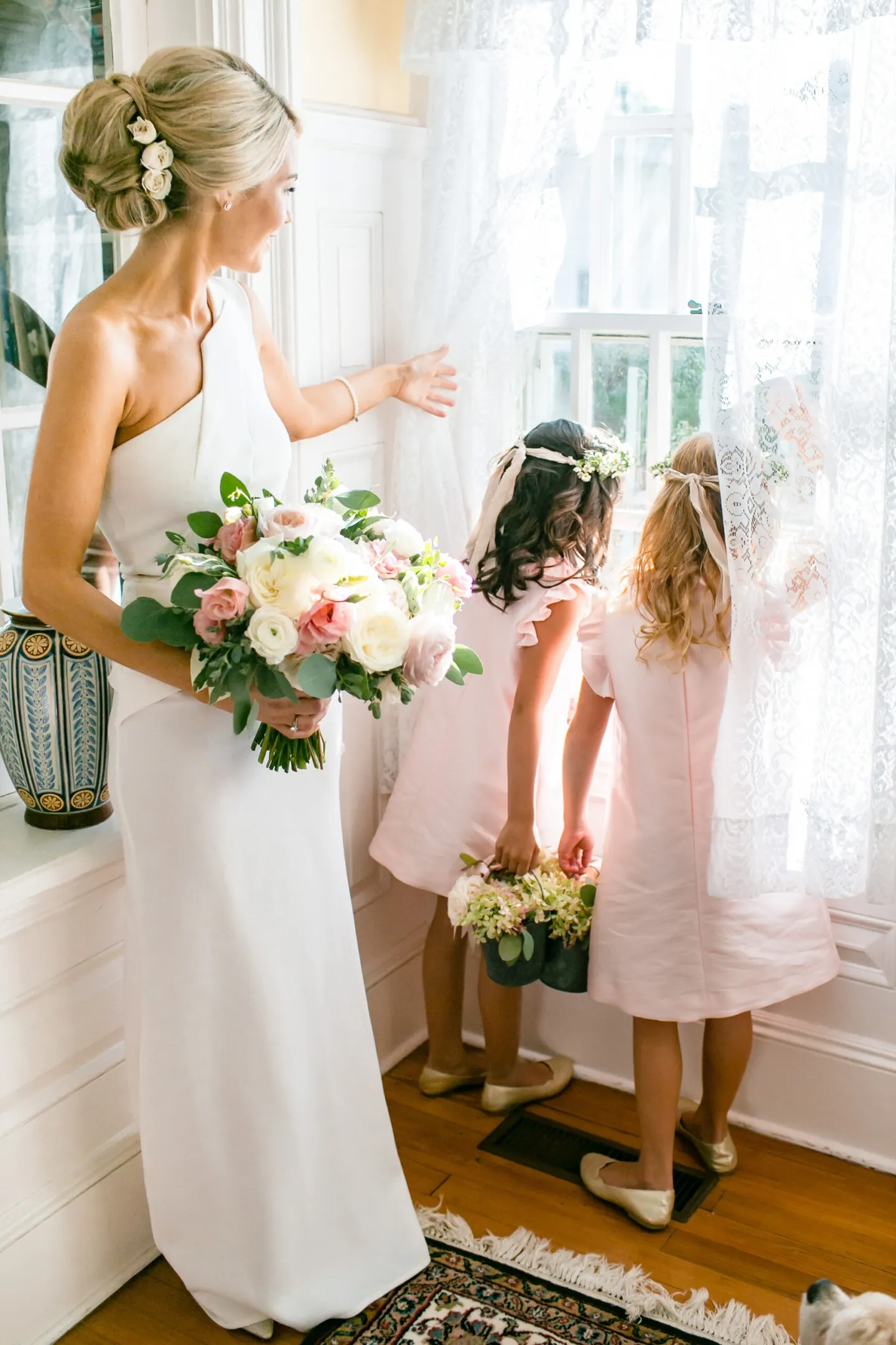 The bride holds her bouquet by a sunny window as flower girls in pink peek outside
