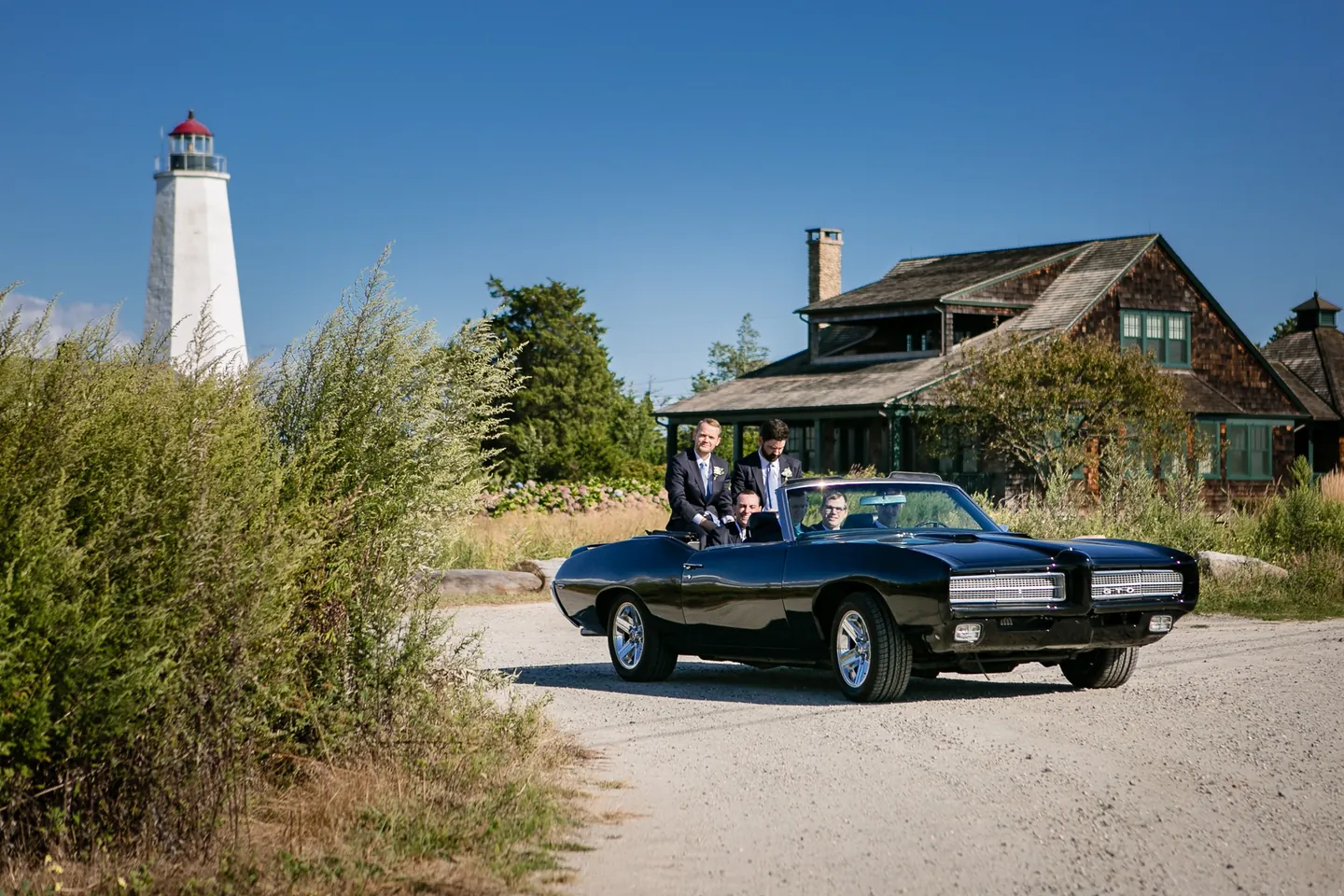 Groomsmen ride in a black vintage convertible along a gravel road below a white lighthouse
