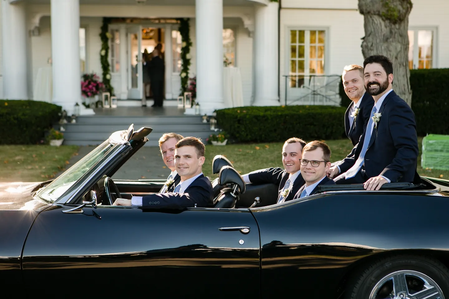 Groomsmen sit up on the back of the black convertible in front of a white-columned house