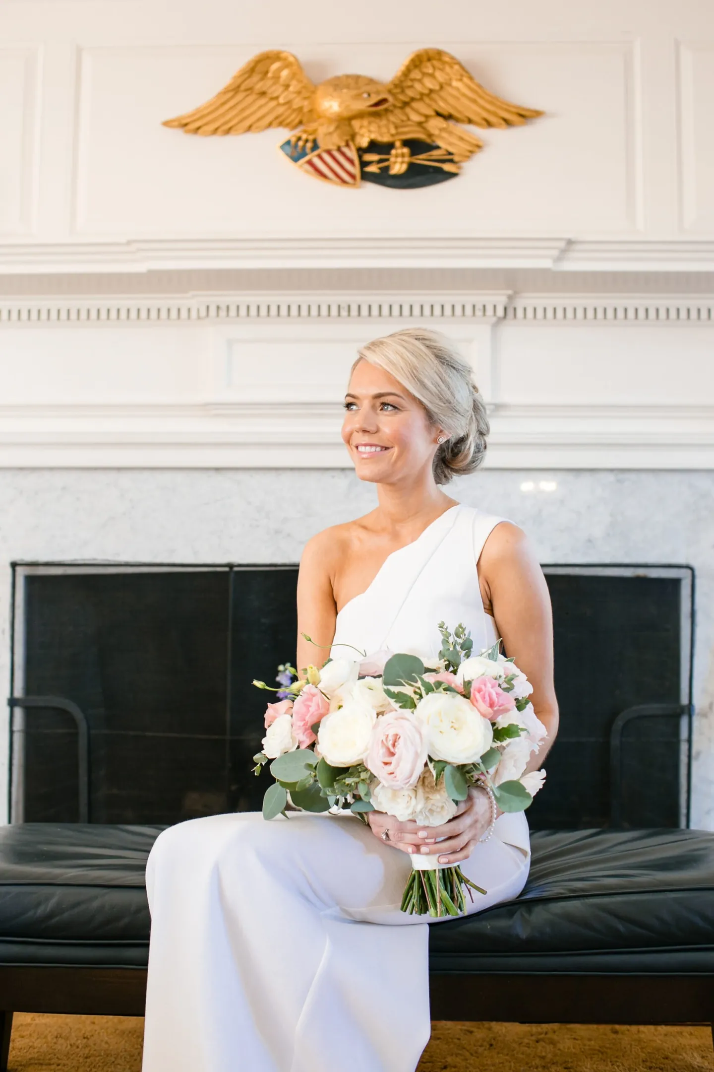 The bride sits on a leather bench below a gilded eagle, holding a pastel bouquet