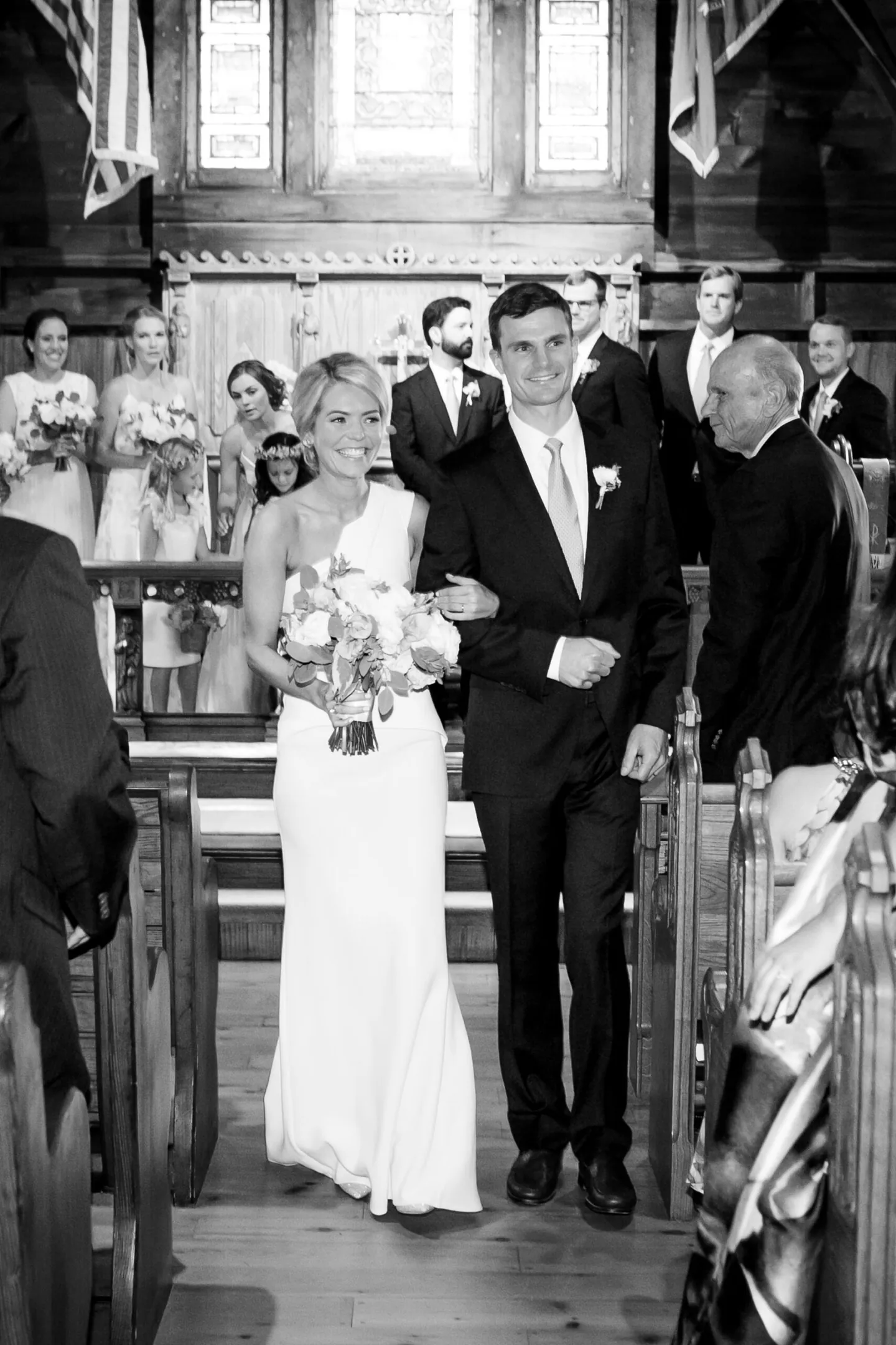 Black and white recessional, the couple walking out of a wooden church past smiling guests