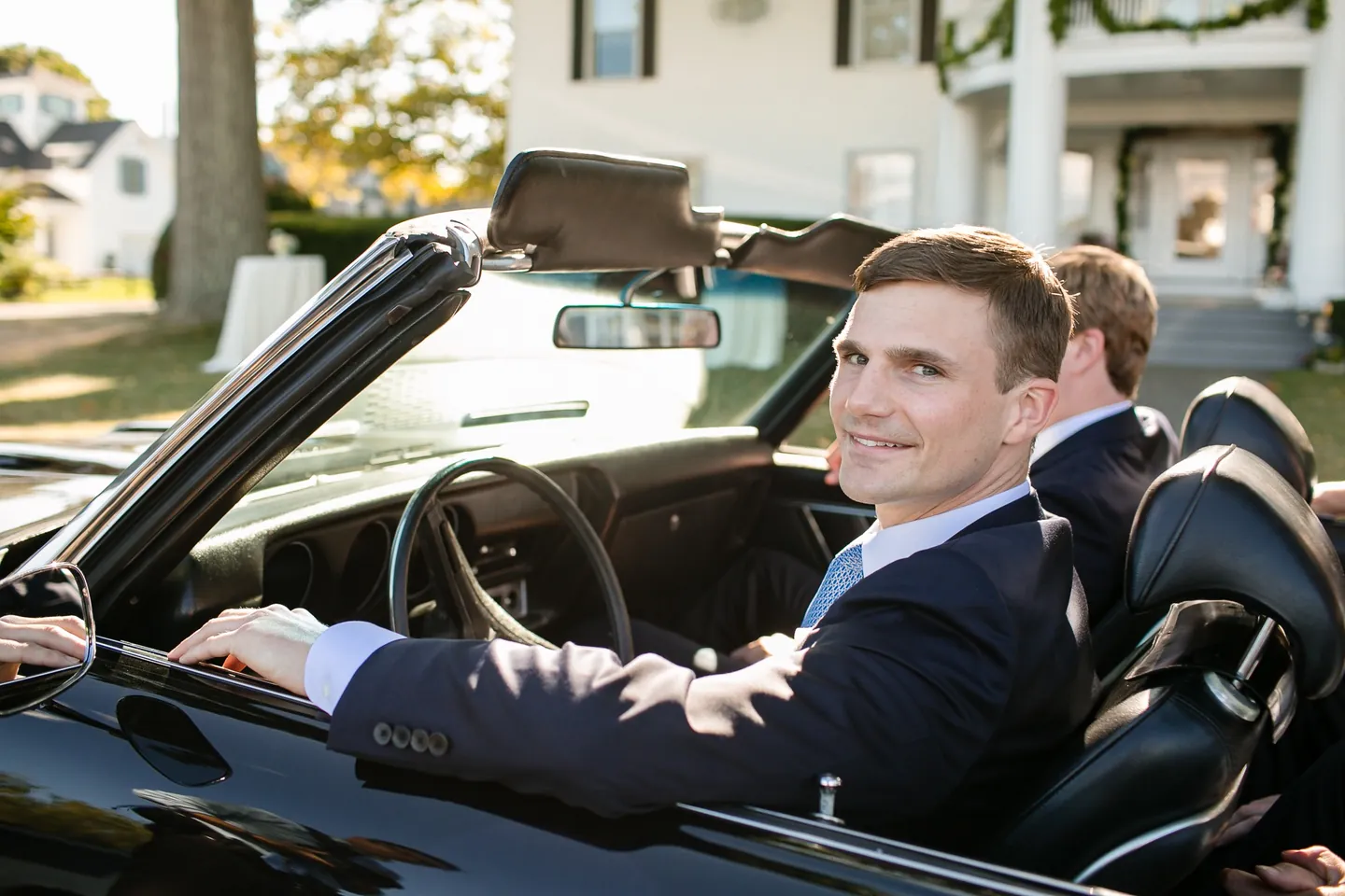 The groom grins behind the wheel of the black convertible outside a white house