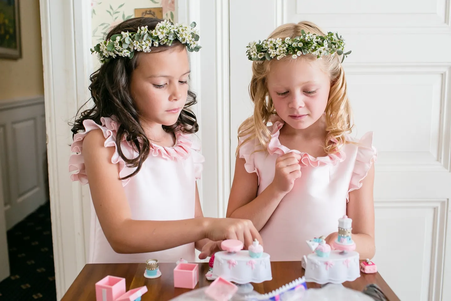 Two flower girls in floral crowns and pink dresses play at a small table