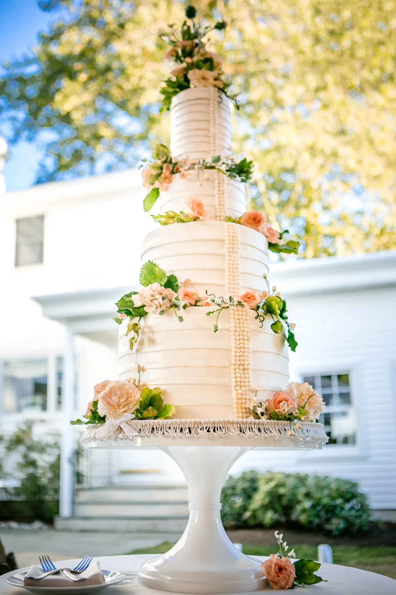 A tall white tiered cake dressed with blush flowers on a stand outdoors