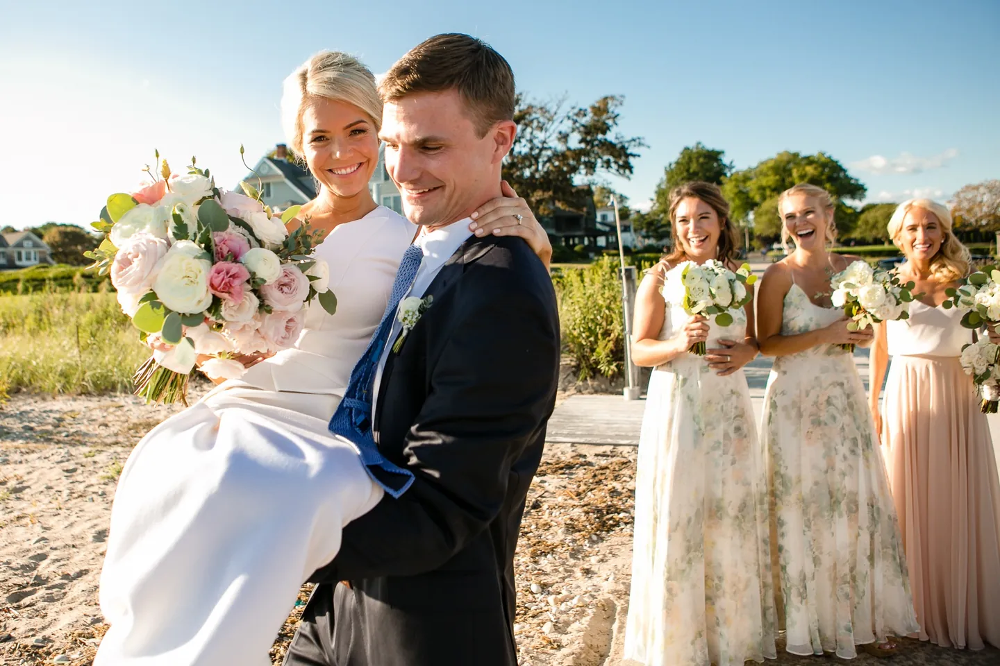 Charlie carries Kristen on the sand while bridesmaids in floral gowns laugh nearby
