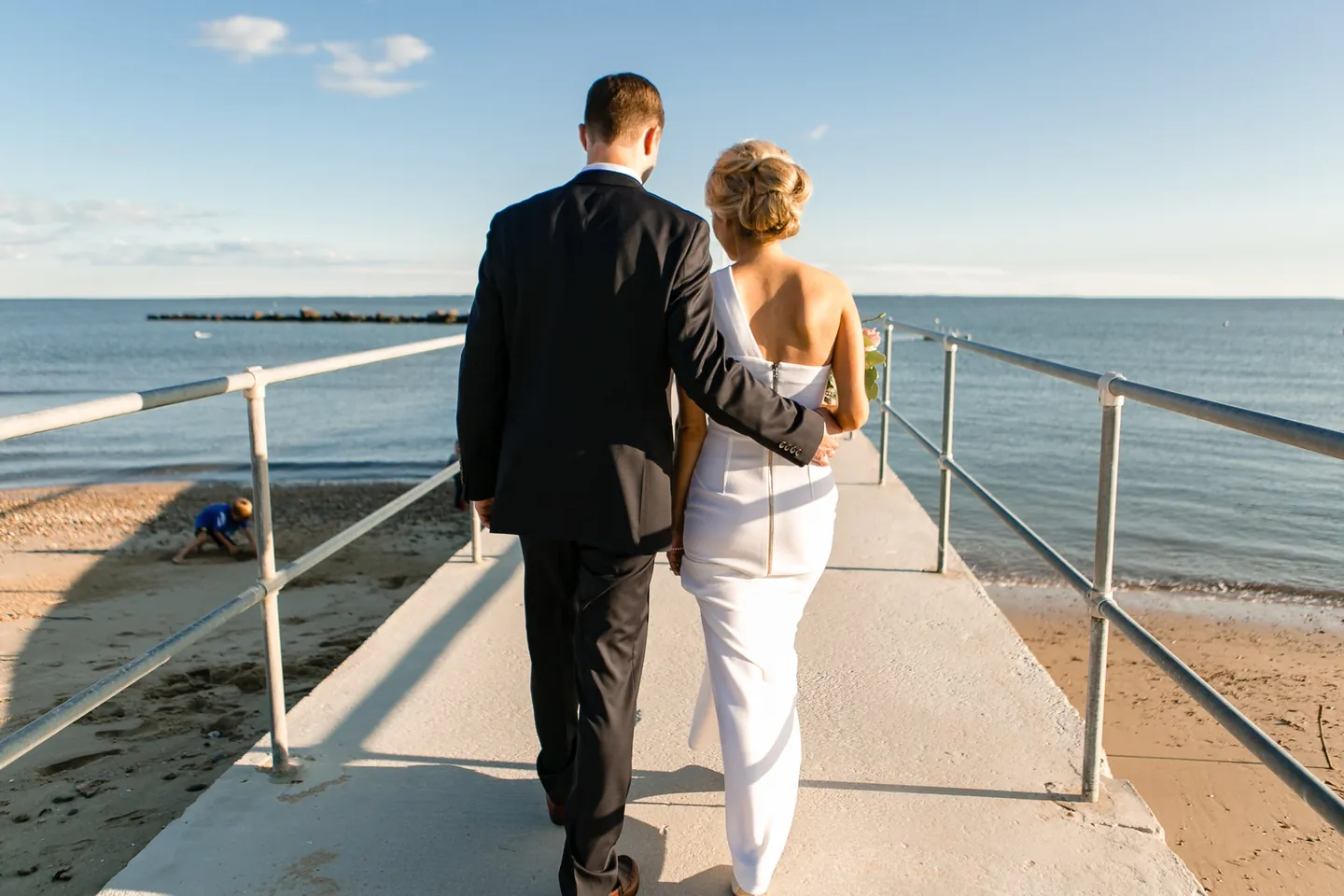 Kristen and Charlie walk arm in arm down a concrete pier toward the open water