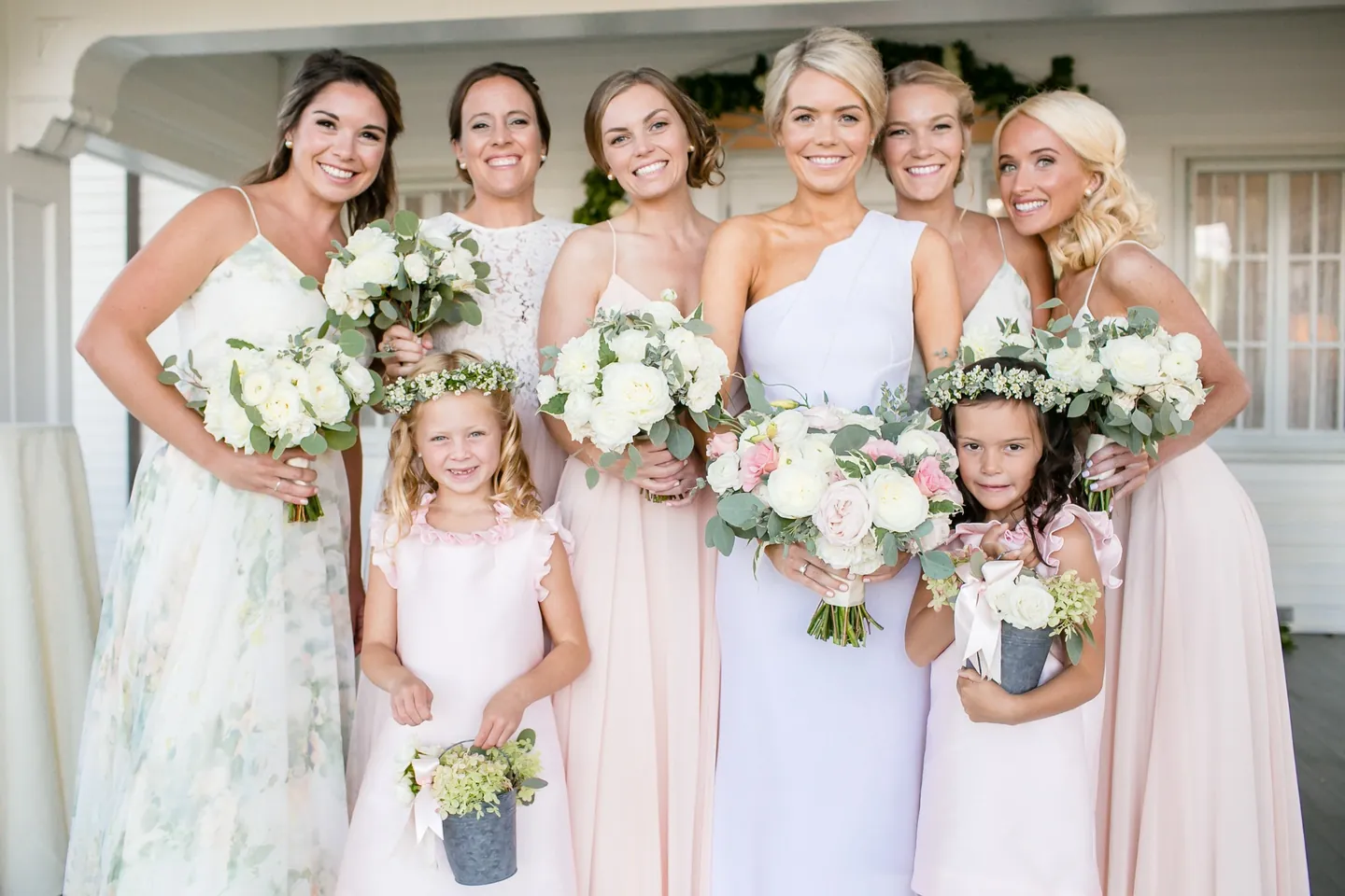 The bride with bridesmaids in blush and floral gowns and 2 flower girls on a porch