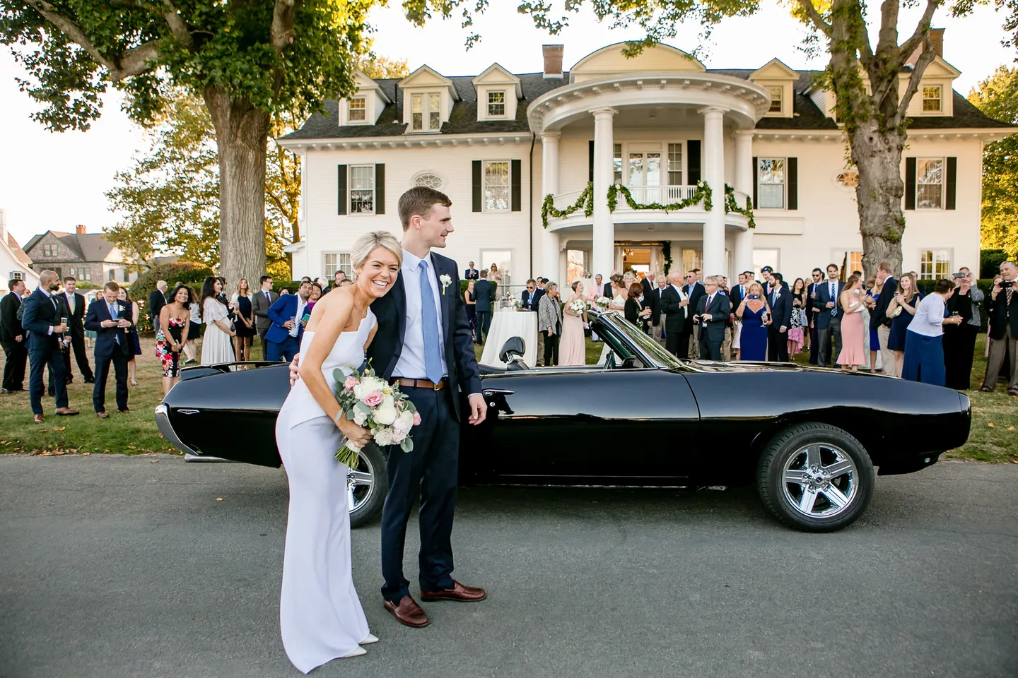 Kristen and Charlie laugh beside the black convertible as guests gather at a white-columned house