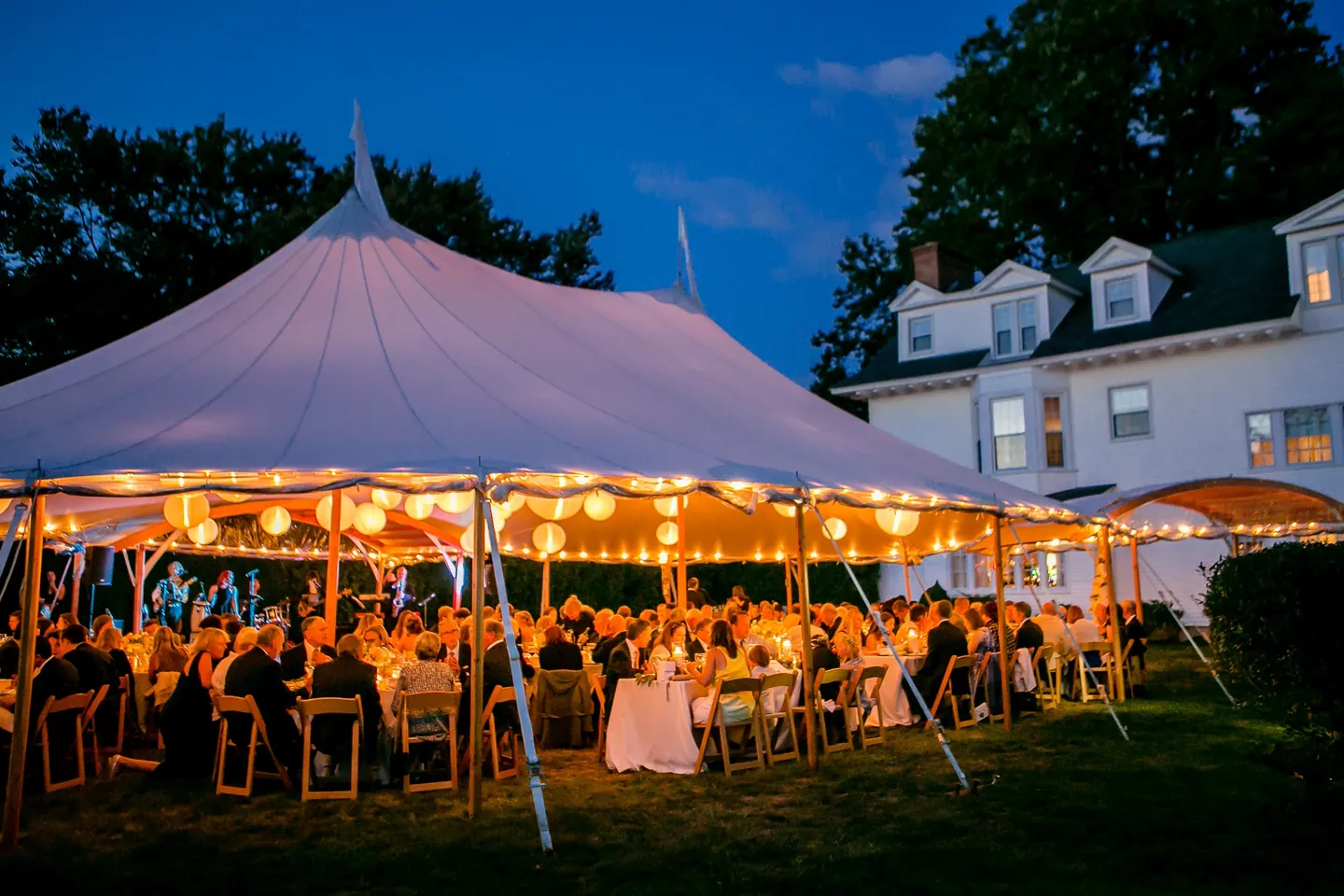 The sailcloth tent glows warm at dusk beside a white house under a blue sky