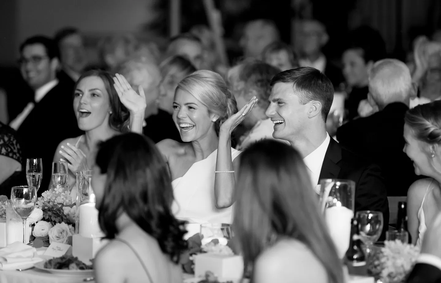 Black and white frame of the bride laughing and waving at her table during toasts