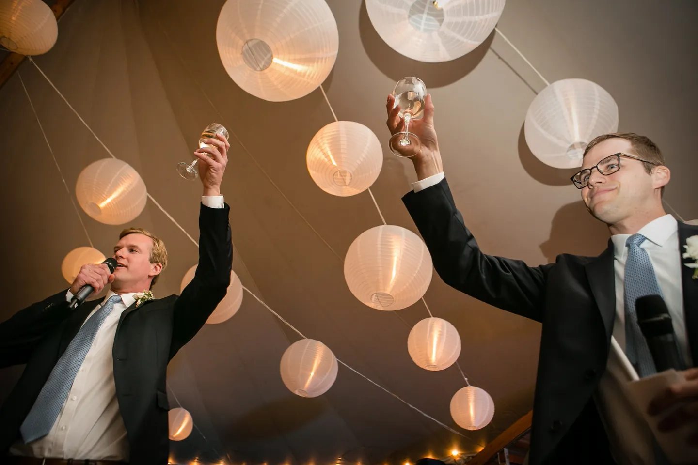 Two men in suits raise champagne glasses high beneath glowing paper lanterns