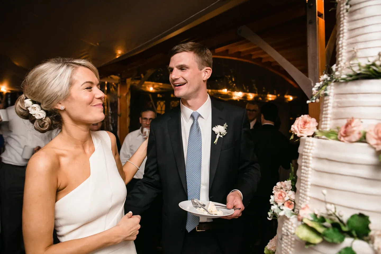 Kristen and Charlie laugh beside the tiered cake holding a plate after cutting it