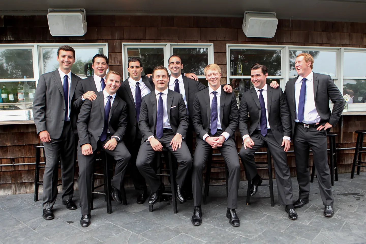 Groomsmen in grey suits and purple ties sit and stand along a shingled patio