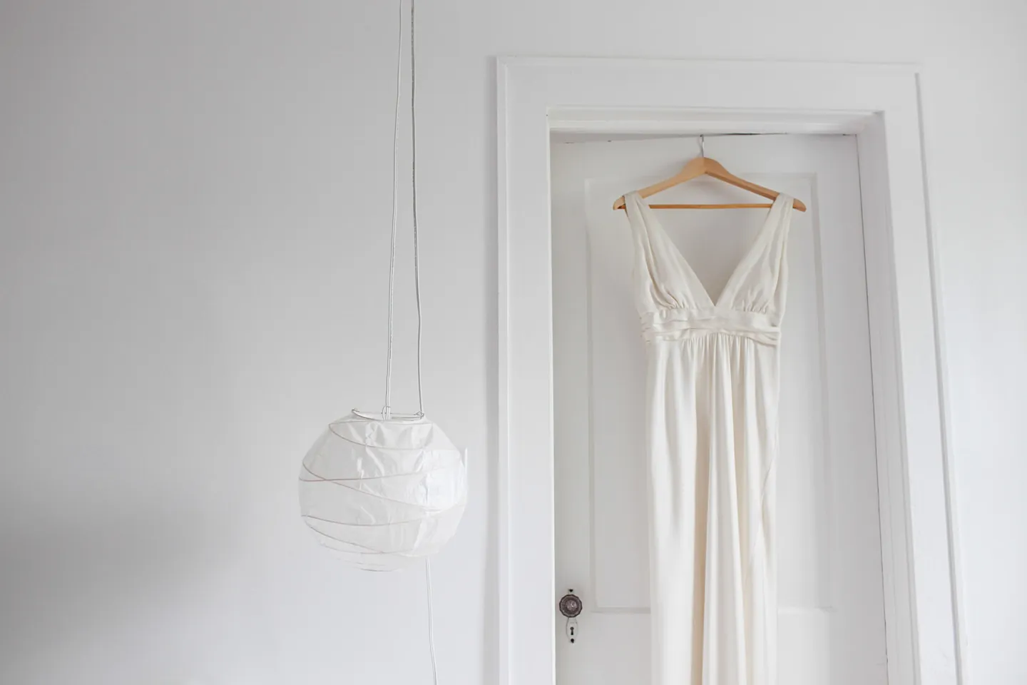 A simple ivory wedding gown hangs from a white door in a bright room