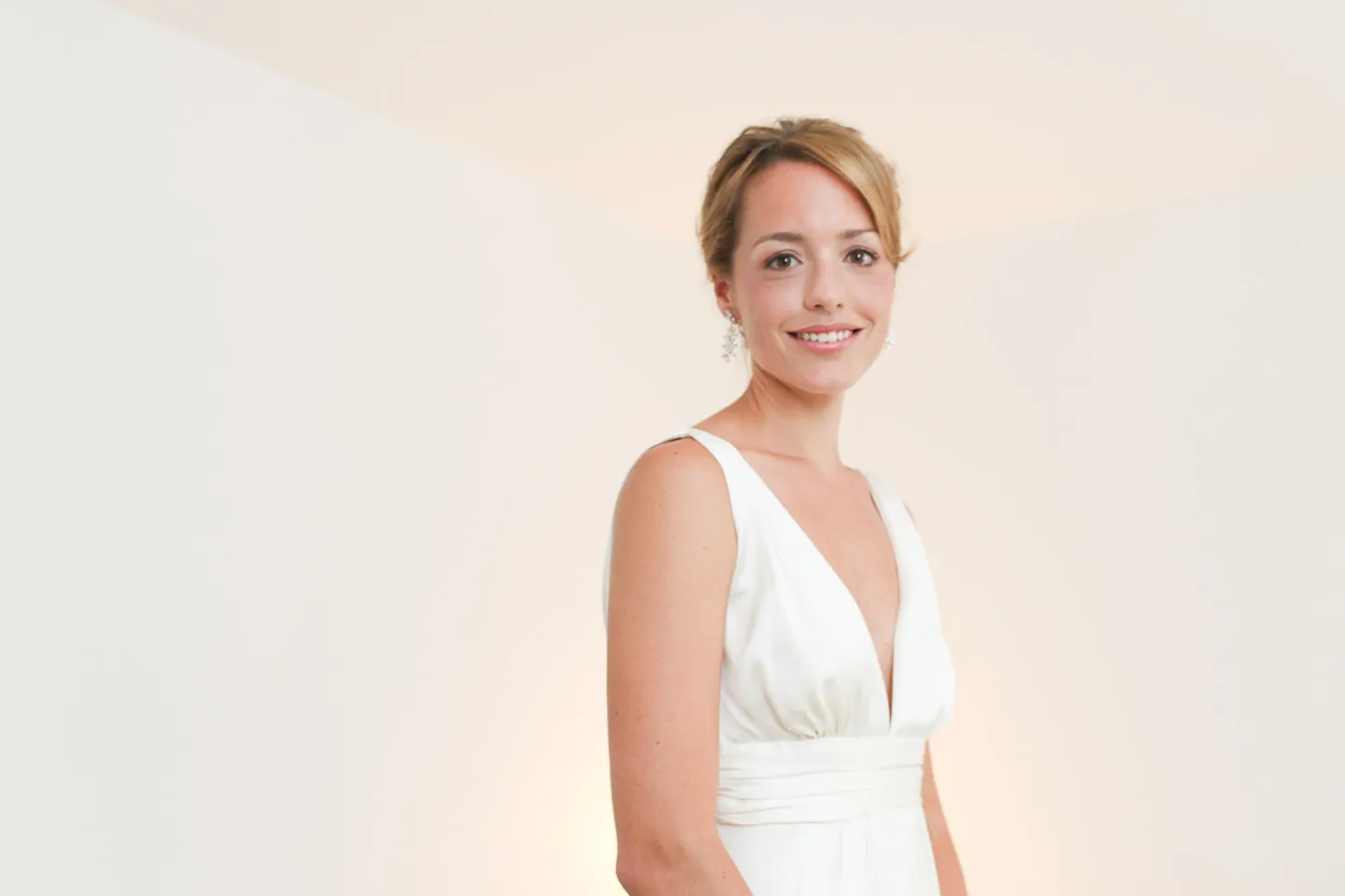 The bride stands in soft window light, hair pinned low, in a V-neck gown